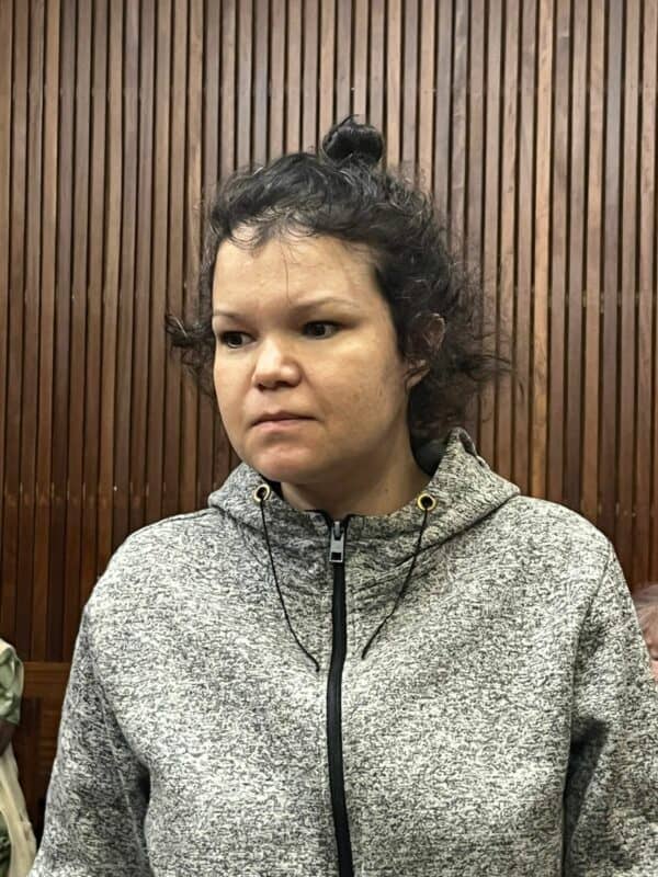 A person with a neutral expression stands in a room with wooden paneling, wearing a gray zip-up hoodie. The mood appears calm and contemplative.