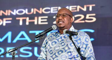 A man stands at a podium speaking into two microphones, wearing glasses and a patterned blue shirt. Behind him, a screen displays bold text.