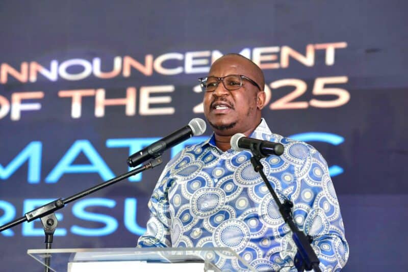 A man stands at a podium speaking into two microphones, wearing glasses and a patterned blue shirt. Behind him, a screen displays bold text.