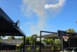 Suburban backyard with a parked white SUV, a basketball hoop, and distant smoke rising against a clear blue sky, under a covered patio.