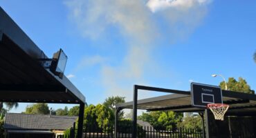 Suburban backyard with a parked white SUV, a basketball hoop, and distant smoke rising against a clear blue sky, under a covered patio.