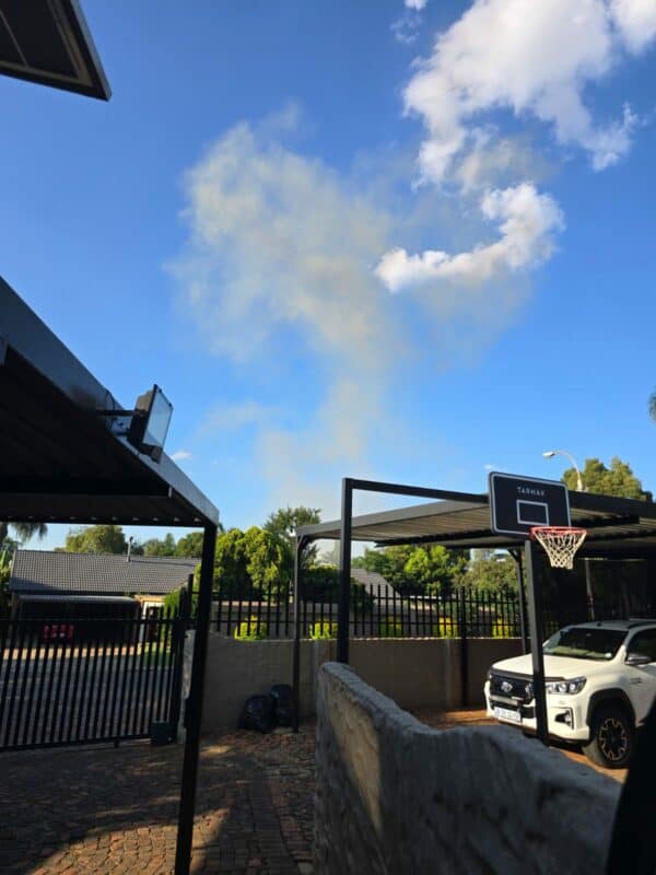 Suburban backyard with a parked white SUV, a basketball hoop, and distant smoke rising against a clear blue sky, under a covered patio.