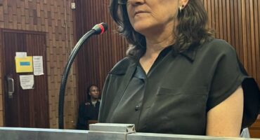 A woman stands in a courtroom, speaking into a microphone. She wears glasses and a black shirt, with a serious expression. Wood paneling surrounds her.