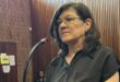 A woman stands in a courtroom, speaking into a microphone. She wears glasses and a black shirt, with a serious expression. Wood paneling surrounds her.
