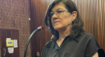 A woman stands in a courtroom, speaking into a microphone. She wears glasses and a black shirt, with a serious expression. Wood paneling surrounds her.