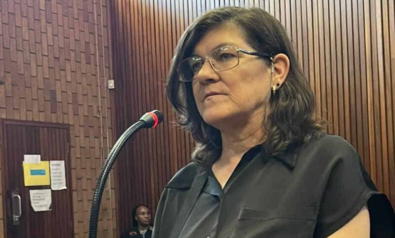 A woman stands in a courtroom, speaking into a microphone. She wears glasses and a black shirt, with a serious expression. Wood paneling surrounds her.