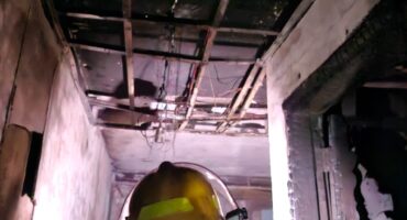 A firefighter wearing a helmet stands in a charred hallway, inspecting a damaged ceiling with exposed beams and wires, capturing a sense of destruction.