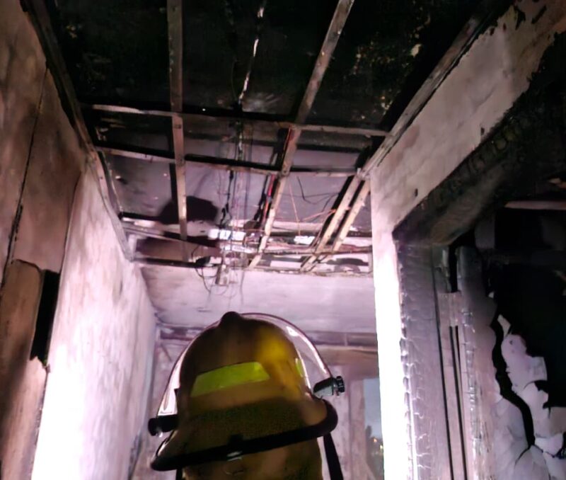 A firefighter wearing a helmet stands in a charred hallway, inspecting a damaged ceiling with exposed beams and wires, capturing a sense of destruction.