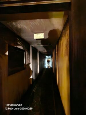 Dimly lit hallway with charred walls, leading to a bright exit. Two firefighters in silhouette are visible at the end. Sign reads “Level 7.” Eerie atmosphere.