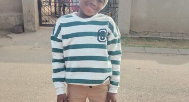 A smiling child stands on a quiet street, wearing a striped sweater, beige pants, and a knit hat. Behind is a gate and sunny sky, creating a cheerful mood.