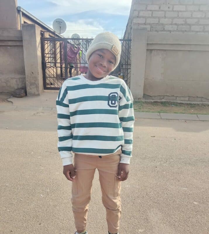 A smiling child stands on a quiet street, wearing a striped sweater, beige pants, and a knit hat. Behind is a gate and sunny sky, creating a cheerful mood.