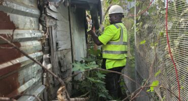 A person in a high-visibility vest and hard hat inspects a weathered corrugated metal structure. They stand between the structure and a fence covered in green vines, conveying a tone of focus in a dense environment.