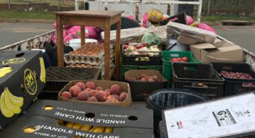 A truck bed filled with assorted goods including boxes of bananas, peaches, egg cartons, and crates. The scene is busy and practical under a cloudy sky.