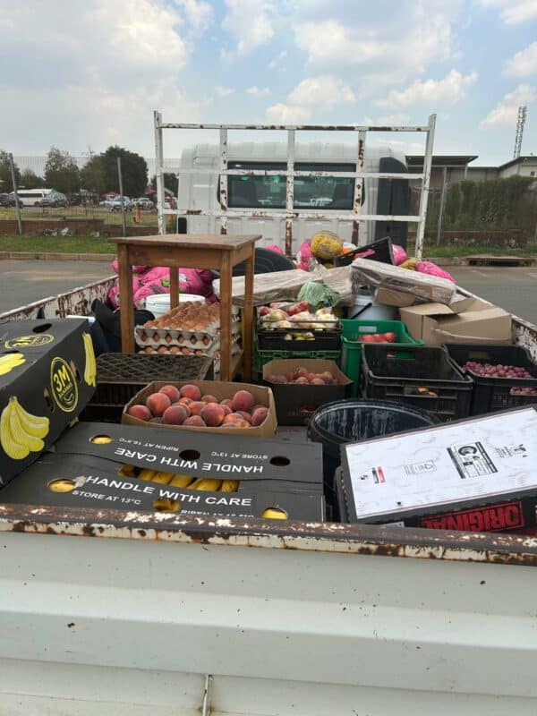 A truck bed filled with assorted goods including boxes of bananas, peaches, egg cartons, and crates. The scene is busy and practical under a cloudy sky.