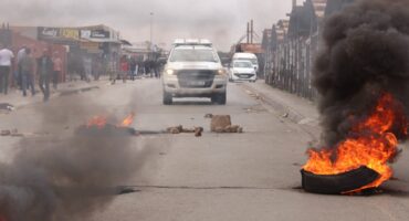 A street scene features burning tires emitting thick black smoke and flames, with a police vehicle advancing. People stand along both sides, creating a tense atmosphere.