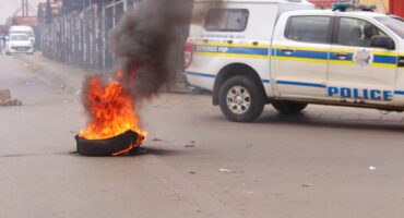 A tire is burning intensely in the middle of a street, emitting thick black smoke. A police vehicle is parked nearby, creating a tense atmosphere