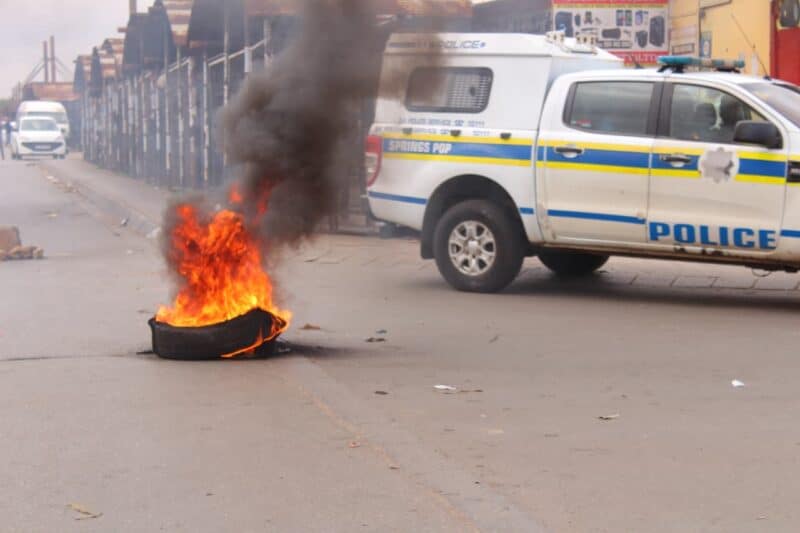 A tire is burning intensely in the middle of a street, emitting thick black smoke. A police vehicle is parked nearby, creating a tense atmosphere