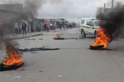 Street scene with burning tires emitting thick smoke, scattered rocks, and a police van nearby. People are gathered on the sidewalk, suggesting unrest.