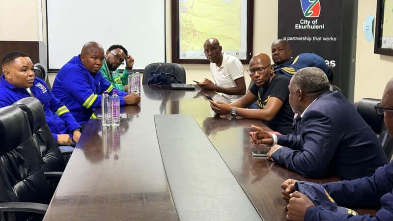 A group of men sit around a conference table in a meeting room, engaged in discussion. Some wear blue uniforms, indicating a collaborative meeting. A city map and banner adorn the walls, setting a professional tone.