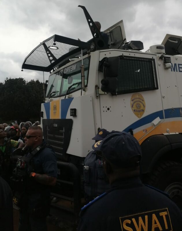 Police officers stand beside an armored vehicle with grilles over its windows, under cloudy skies. The atmosphere is tense, with a crowd nearby.