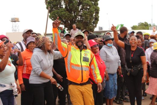 An angry crowd of people stands together outdoors, with a man in a bright orange jacket raising his arm enthusiastically. 