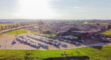Aerial view of a large parking area filled with cars next to a cluster of buildings under a bright, sunny sky. Green grass and a few people are in the foreground.