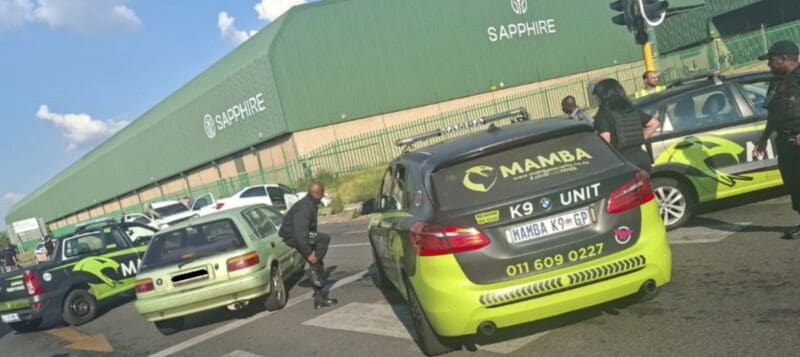Security vehicles and personnel are gathered at an intersection near a large green building labeled