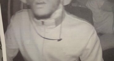 A grainy black-and-white photo shows a person wearing a light jacket and bucket hat. The individual appears surprised, with another figure blurred in the background.