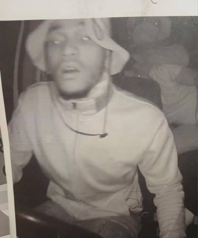 A grainy black-and-white photo shows a person wearing a light jacket and bucket hat. The individual appears surprised, with another figure blurred in the background.