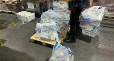 A worker stands in a warehouse beside a pallet covered with large sealed bags filled with packaged items. The scene is industrial and busy.