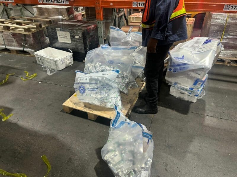 A worker stands in a warehouse beside a pallet covered with large sealed bags filled with packaged items. The scene is industrial and busy.