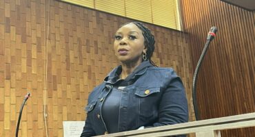 A woman in a blue outfit stands confidently at a courtroom lectern, surrounded by wood paneling. The mood is serious and professional.
