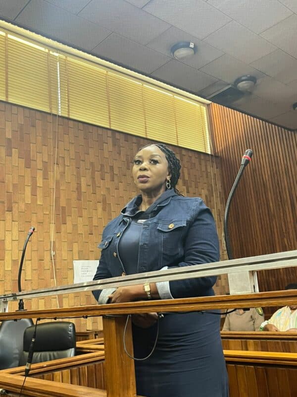 A woman in a blue outfit stands confidently at a courtroom lectern, surrounded by wood paneling. The mood is serious and professional.
