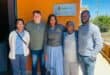 A diverse group of five people smiling outside a building with an "AfriForum Ontvangs" sign on an orange wall. They exude a positive and friendly atmosphere.