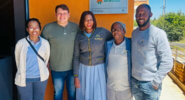 A diverse group of five people smiling outside a building with an "AfriForum Ontvangs" sign on an orange wall. They exude a positive and friendly atmosphere.