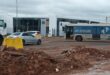 A construction site with loose dirt and rubble in the foreground. Behind, a silver car and a bus with "Turn traffic jams into jam sessions" are on a road. A Volkswagen dealership is in the background under a cloudy sky.