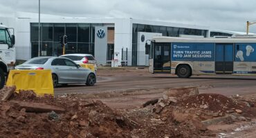 A construction site with loose dirt and rubble in the foreground. Behind, a silver car and a bus with "Turn traffic jams into jam sessions" are on a road. A Volkswagen dealership is in the background under a cloudy sky.