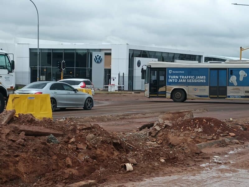 A construction site with loose dirt and rubble in the foreground. Behind, a silver car and a bus with