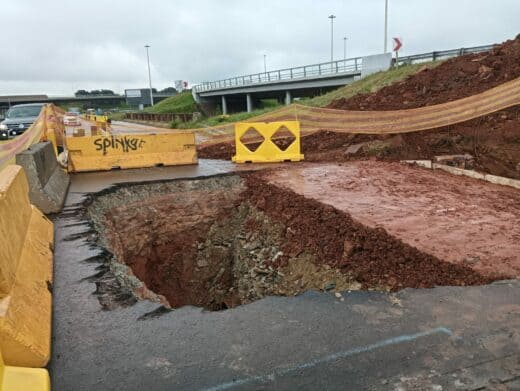 Road construction site with a deep excavation surrounded by yellow barriers and caution tape. Overcast sky, bridge, and cars in the background.
