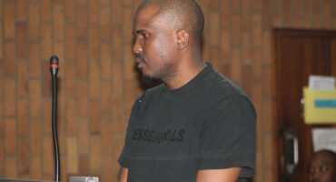 A man wearing a black "ESSENTIALS" t-shirt stands in a courtroom, facing sideways near a microphone. The setting conveys a serious mood.