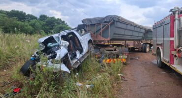 A heavily damaged white car is crushed against a large truck on a dirt road. A fire truck is nearby. The scene is tense, with overcast skies.