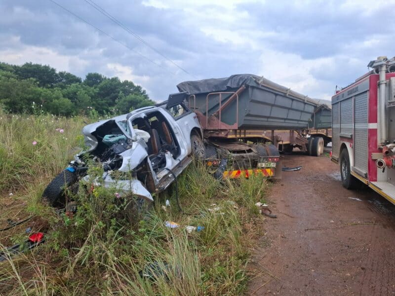 A heavily damaged white car is crushed against a large truck on a dirt road. A fire truck is nearby. The scene is tense, with overcast skies.