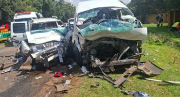 Head-on collision between a white van and SUV on a road. Both vehicles are severely damaged. Debris is scattered, with emergency responders nearby.