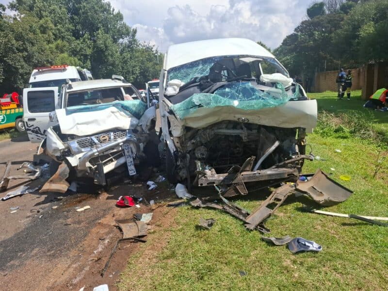 Head-on collision between a white van and SUV on a road. Both vehicles are severely damaged. Debris is scattered, with emergency responders nearby.