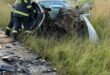 Firefighters in protective gear assess a severely damaged car stranded in tall grass by a roadside, surrounded by debris. Scene conveys urgency.