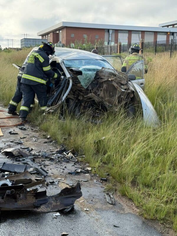 Firefighters in protective gear assess a severely damaged car stranded in tall grass by a roadside, surrounded by debris. Scene conveys urgency.