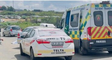 Ambulance and private security car on a road, with two people in uniform conversing nearby. The scene is set in a semi-rural area under a partly cloudy sky.