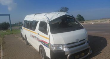 A white van with significant roof damage is parked on a roadside. The vehicle has colorful stripes along the side. The sky is clear and blue.