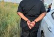 A person in all-black clothing is handcuffed beside a white car on a road. Grass and fencing appear on the left. The scene suggests an arrest.