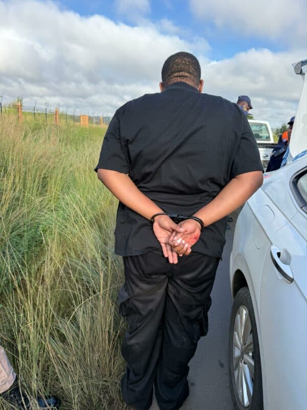 A person in all-black clothing is handcuffed beside a white car on a road. Grass and fencing appear on the left. The scene suggests an arrest.